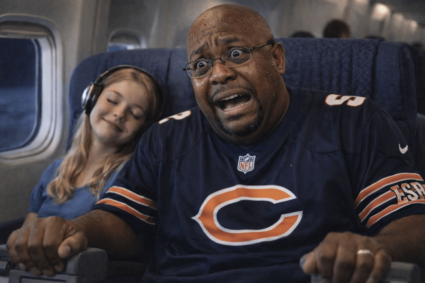An image of a 30-year-old black man wearing a Chicago Bears jersey. He is sitting on a plane next to a young white girl. He is panicking as the plane flies through turbulence. She is fast asleep and enjoying the flight.